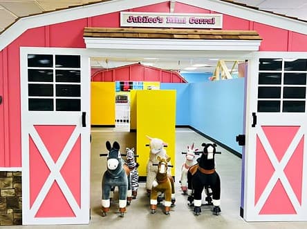 Jubilee's Mini Corral, a pink barn with open doors and plush ride-on horses, a zebra and a llama inside