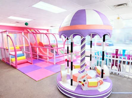 Lavender and white soft play carousel with padded animal seats, in front of the pink soft play climbing structure