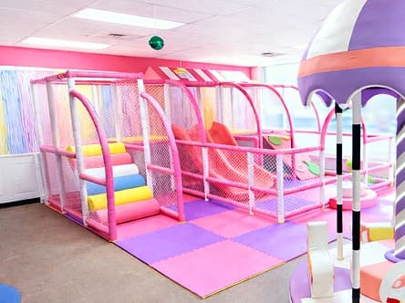 Pink soft play structure with padded steps, netted climbing levels and slides over purple and pink floor mats